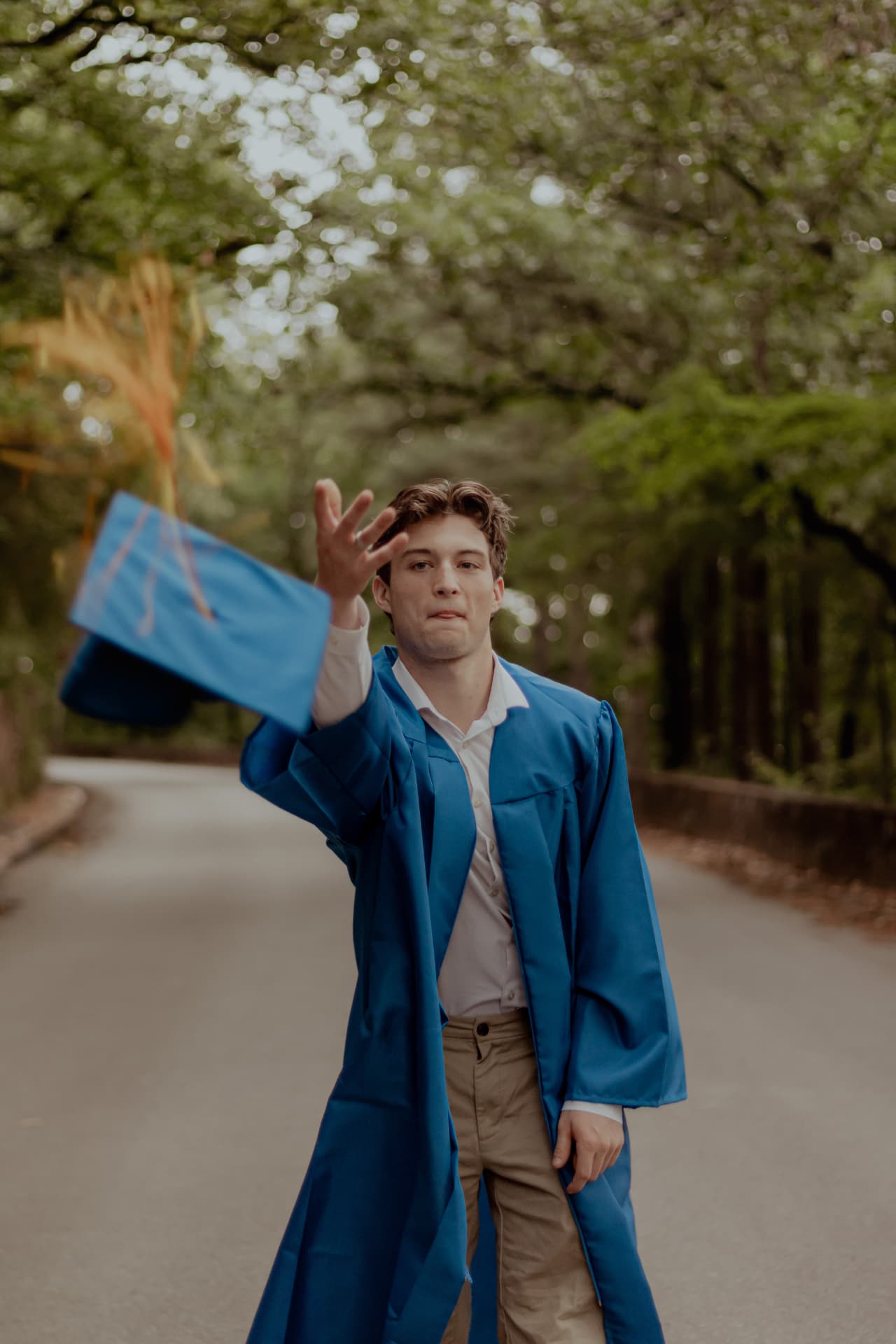 Young man tossing his graduation cap into the air on a tree-lined road